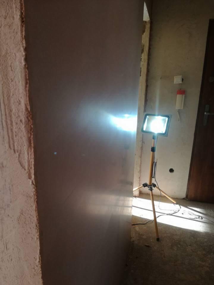Świeżo otynkowana ściana z widocznym punktowym oświetleniem roboczym na drewnianym trójnogu, nierówna powierzchnia ściany przed otynkowaniem po lewej stronie.