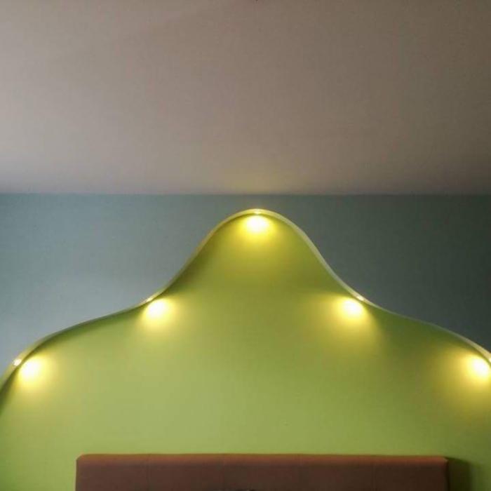 Nietypowe wezgłowie łóżka w kształcie fali, pomalowane na jasnozielony kolor, z wbudowanymi punktowymi lampkami LED dającymi ciepłe światło, na tle ściany w kolorze morskim i białego sufitu.