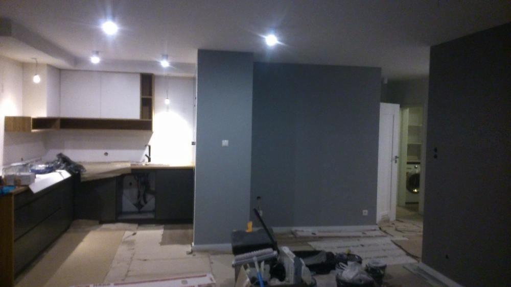 Wnętrze w trakcie prac wykończeniowych: szare ściany, częściowo zamontowana zabudowa kuchenna z drewnianymi półkami, podłoga zabezpieczona kartonem, widoczne narzędzia malarskie i sprzęt AGD w tle.