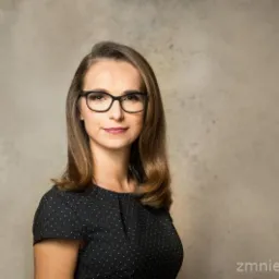 Portret kobiety w okularach, ubranej w czarną sukienkę w białe kropki, na neutralnym tle.