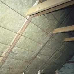 Izolacja poddasza z widocznymi drewnianymi belkami i drutem mocującym, jasna wełna mineralna między krokwiami, fragment muru z oknem w tle.