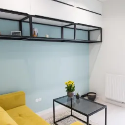 Salon z żółtą sofą, szklanym stolikiem kawowym z kwiatami i czarnymi, metalowymi regałami ściennymi nad turkusową ścianą.