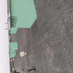 Widok z góry na płaski dach budynku z pokryciem bitumicznym, fragmentem pomalowanej na zielono attyki i elementami wentylacji, w tle zabudowa miejska przy pochmurnym niebie.