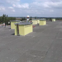 Widok z góry na płaski dach pokryty papą z żółtymi, świeżo pomalowanymi obudowami wentylacyjnymi; w tle budynki i drzewa pod błękitnym niebem z chmurami.