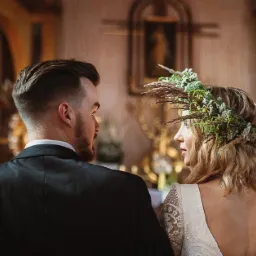 Para młoda stoi przed ołtarzem w kościele, panna młoda ma wianek z zielonych gałązek i suszonych traw, a pan młody ma na sobie czarny garnitur.