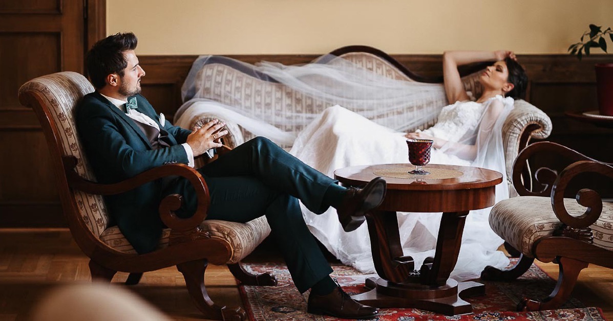 Elegancki pan młody w zielonym garniturze siedzi w fotelu, podczas gdy panna młoda w białej sukni leży na kanapie w tle, na stoliku stoi kieliszek.