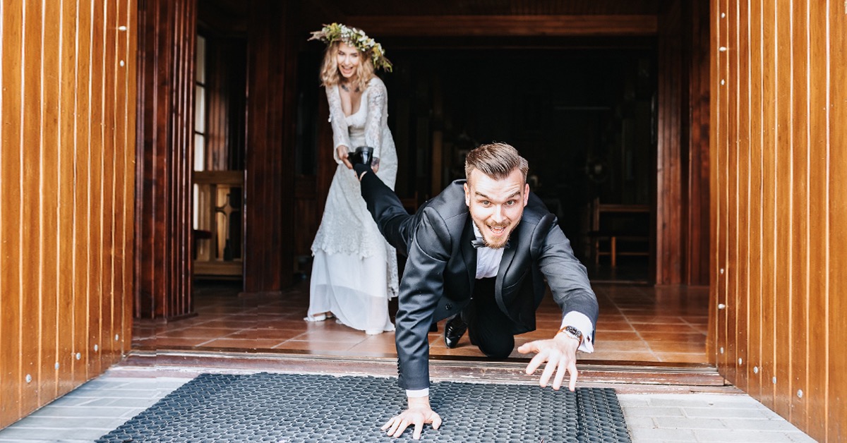 Panna młoda trzyma but pana młodego, który na kolanach wchodzi do drewnianego budynku z szerokim uśmiechem.