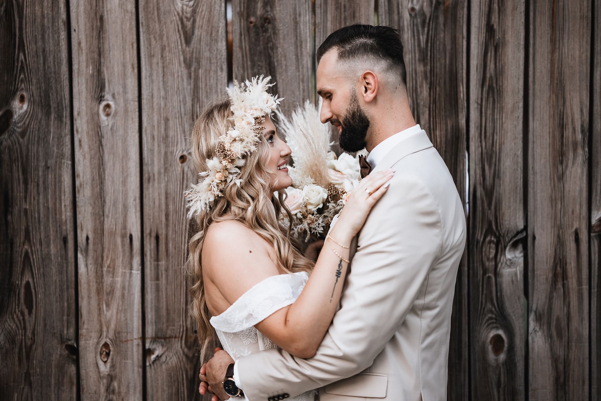 Uśmiechnięta para młoda w beżowych strojach, obejmująca się na tle drewnianej ściany; panna młoda ma wianek z suszonych kwiatów i delikatny bukiet, widoczny tatuaż na jej ramieniu.