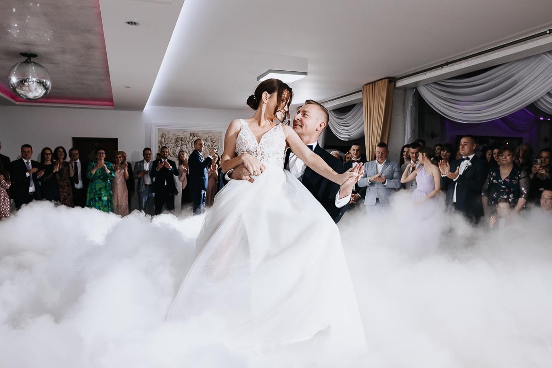 Pierwszy taniec pary młodej w chmurach dymu na tle gości weselnych, suknia ślubna z koronkową górą i tiulowym dołem.
