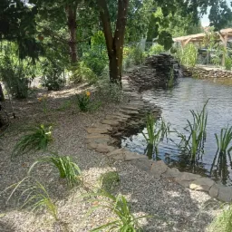 Oczko wodne z kamiennym wodospadem, otoczone roślinnością i ścieżką z płaskich kamieni. Drzewo z lampkami dodaje uroku. W tle widoczna altana.