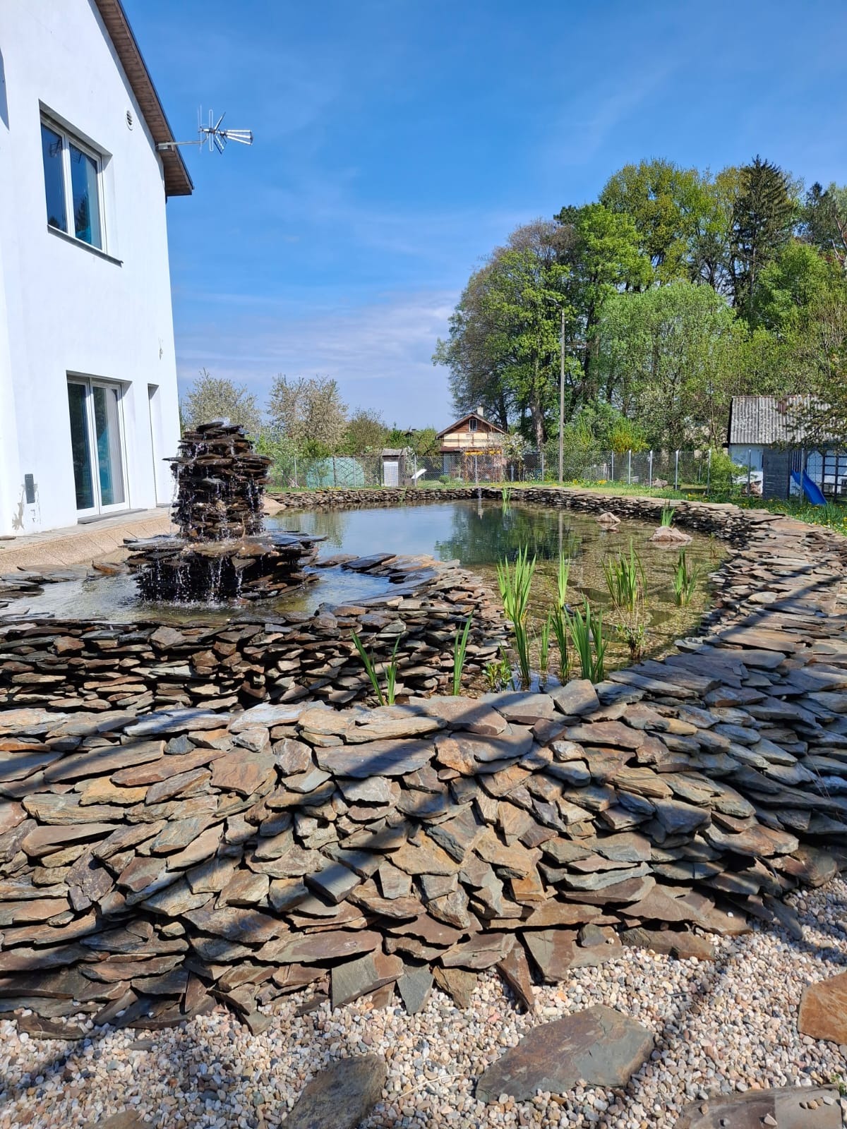 Staw rekreacyjny z kaskadą wodną, obłożony kamieniem łupkowym, otoczony ozdobnym żwirem. W tle dom z białymi ścianami i zielone drzewa.