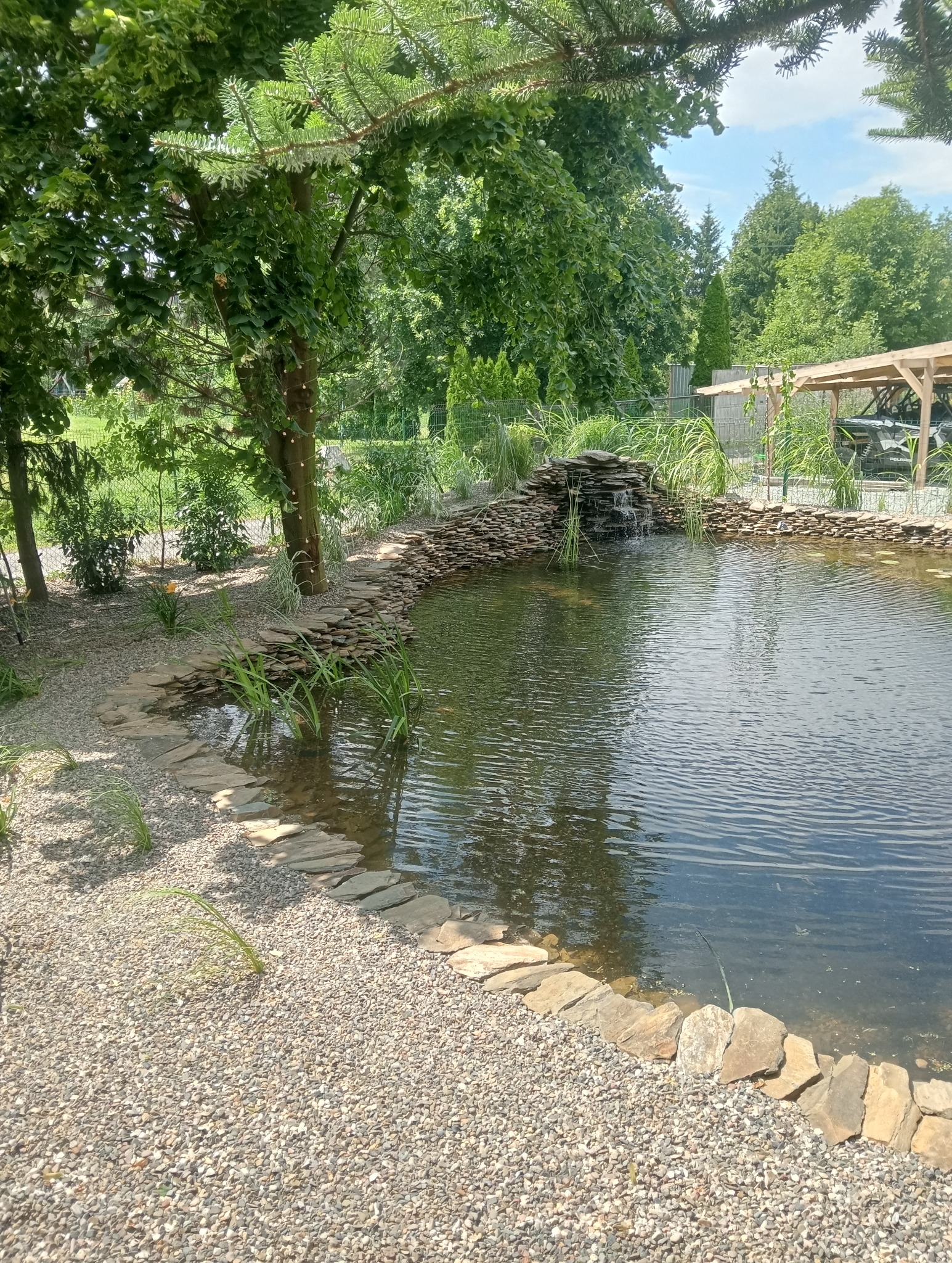 Oczko wodne z kaskadą, otoczone kamieniami i zielenią. Widoczne rośliny wodne i ozdobne. W tle drzewa i krzewy, oraz altana.