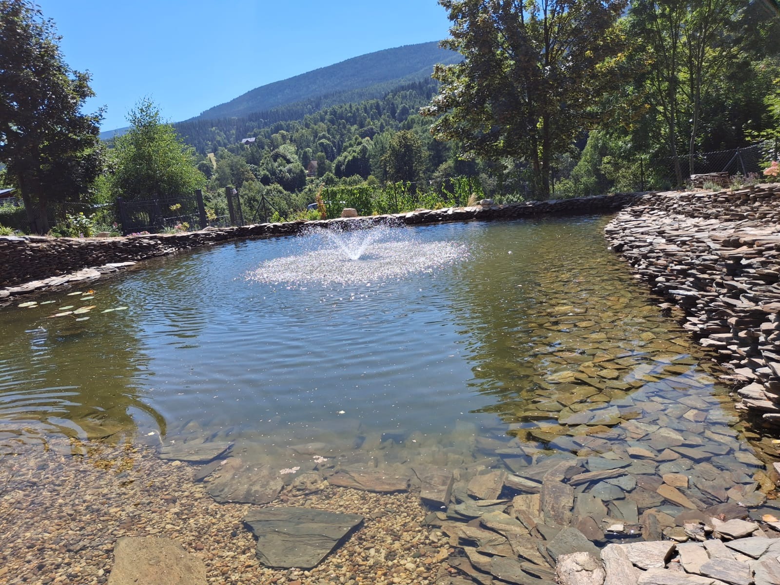 Oczko wodne z fontanną i kamiennym brzegiem na tle zalesionych wzgórz. Woda czysta, z widocznym dnem. Kamienny brzeg z ułożonych płaskich kamieni.