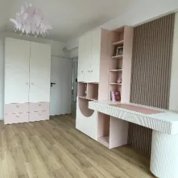 Pokój dziecięcy z białymi szafami i biurkiem na wymiar, akcenty różowe, lampa motylkowa, drewniana podłoga. Całość w jasnych, pastelowych barwach.