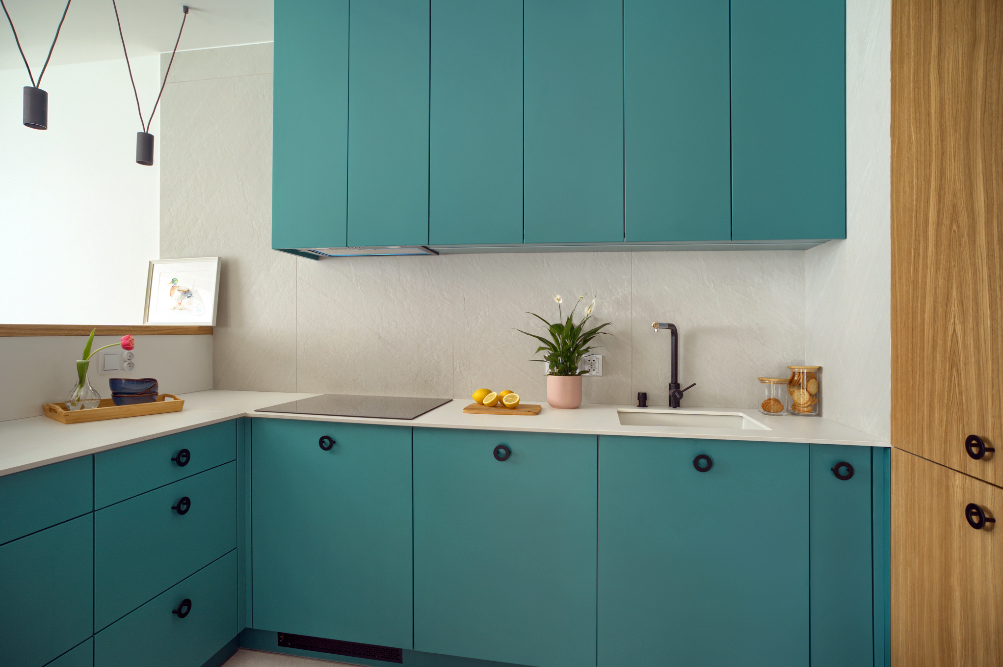 Turkusowa kuchnia na wymiar z czarną armaturą i okrągłymi uchwytami, na blacie deska z cytrynami oraz doniczka z kwiatem, nad blatem wiszące szafki w tym samym kolorze, obok drewniana szafa...