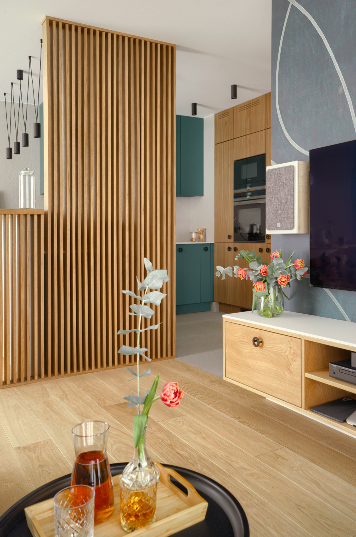 Nowoczesne wnętrze mieszkania z drewnianą ścianką działową, szafkami kuchennymi w odcieniach drewna i zieleni, oraz dekoracjami kwiatowymi i napojami na stoliku kawowym.