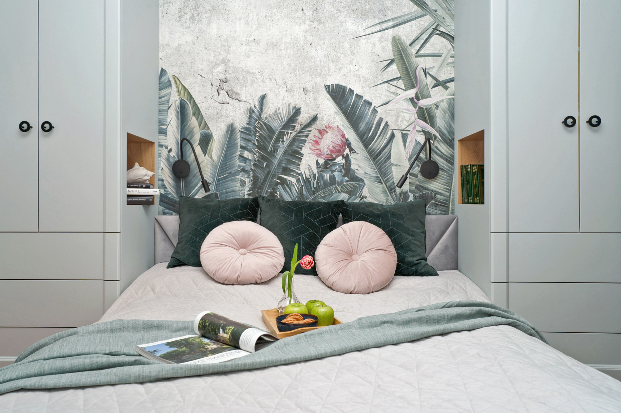 Jasnoszara sypialnia z łóżkiem w zabudowie szafy, tapeta z motywem tropikalnych liści, okrągłe różowe poduszki i śniadanie na tacy.