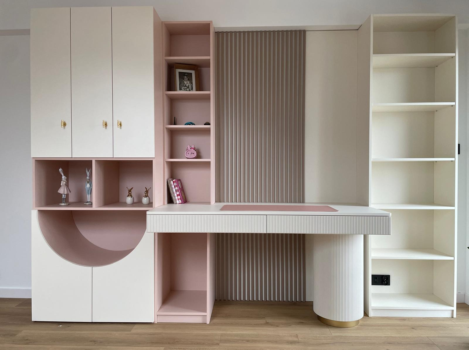 Biało-różowe meble na wymiar: biurko z szafkami, półkami i ryflowanym panelem. Uwagę zwraca półokrągła szafka z dekoracjami. Nowoczesny design.