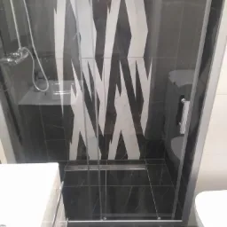 Wykończona łazienka z czarno-białymi płytkami w geometryczne wzory, kabina prysznicowa ze szklanymi drzwiami i odpływem liniowym w podłodze, widoczna umywalka i toaleta