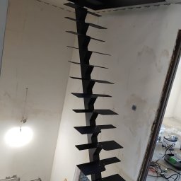 TOTAL METAL - Czarne, metalowe schody wspornikowe z kwadratowymi stopniami, zamontowane na ścianie w surowym wnętrzu podczas remontu, widoczne kable i narzędzia.