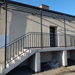 TOTAL METAL - Zewnętrzne schody betonowe z czarną metalową balustradą i zadaszeniem, prowadzące do drzwi wejściowych domu. Widoczne ślady prac budowlanych w otoczeniu.