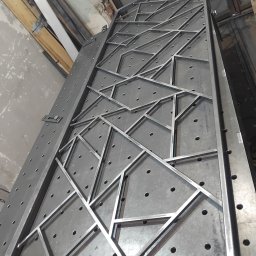 TOTAL METAL - Metalowa konstrukcja z profili o nieregularnych kształtach geometrycznych, spawana na perforowanym stole spawalniczym, widoczne ślady obróbki i surowy stan wykończenia.