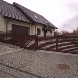 Nowoczesna brama przesuwna z poziomych profili w kolorze brązowym, dopasowana do elewacji domu z brązową stolarką okienną i bramą garażową, widoczna kostka brukowa na podjeździe.