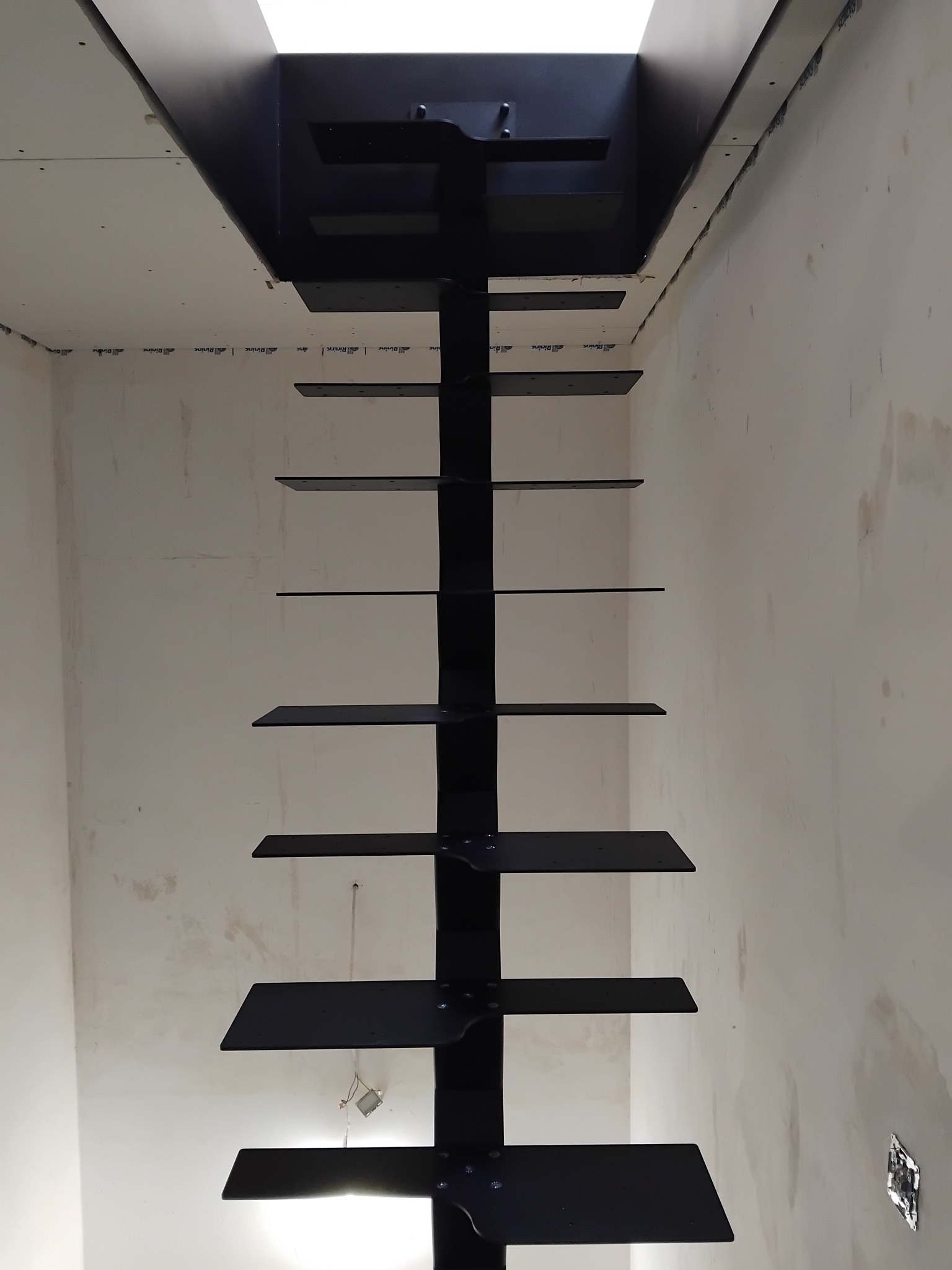 Nowoczesne, minimalistyczne schody metalowe w kolorze czarnym, zamontowane w surowym wnętrzu z widocznymi śladami prac wykończeniowych na ścianach.