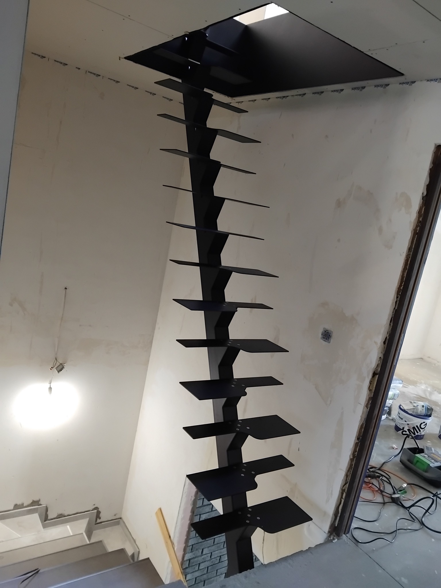 Czarne, metalowe schody wspornikowe z kwadratowymi stopniami, zamontowane na ścianie w surowym wnętrzu podczas remontu, widoczne kable i narzędzia.