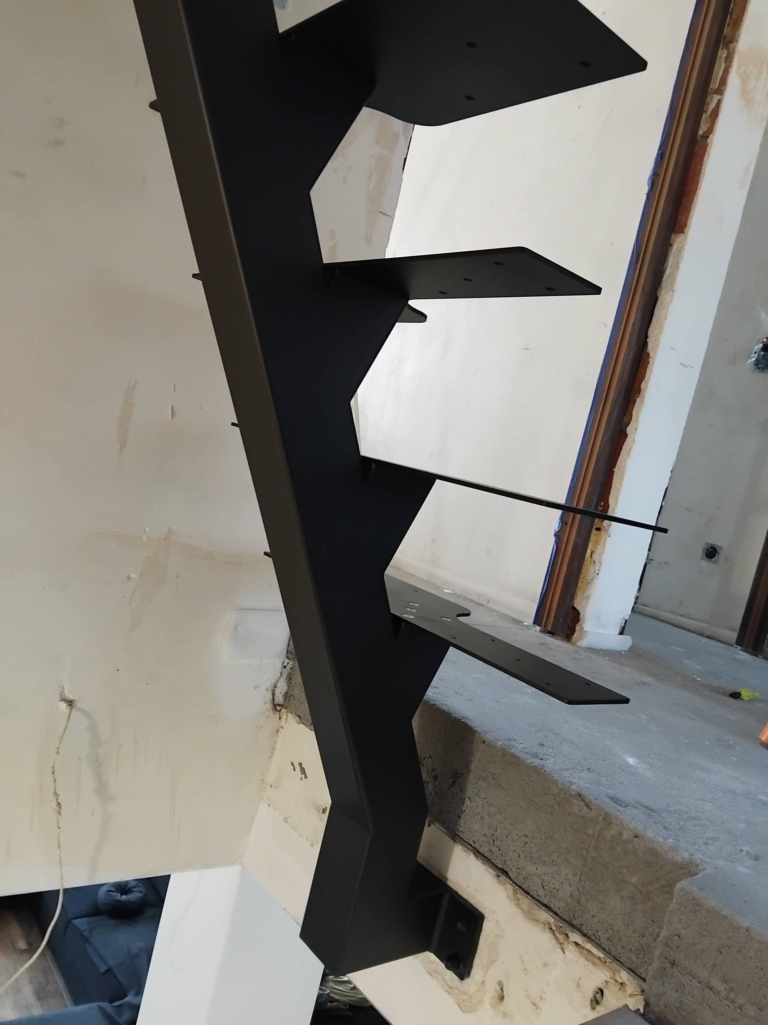 Czarne, metalowe schody wspornikowe z widocznymi mocowaniami do ściany, w surowym stanie wykończenia wnętrza.