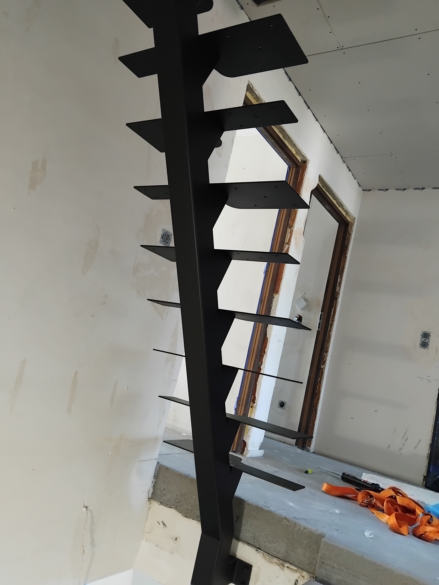 Surowe, metalowe schody policzkowe w trakcie montażu, z widocznymi uchwytami stopni i elementami mocującymi, na tle niewykończonego wnętrza z widocznymi przewodami elektrycznymi i narzędziami.