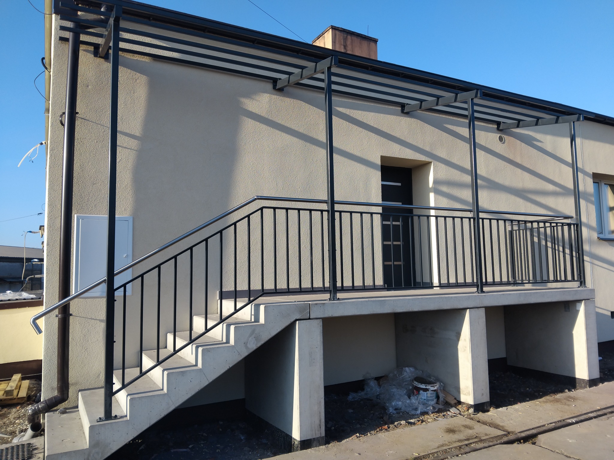 Zewnętrzne schody betonowe z czarną metalową balustradą i zadaszeniem, prowadzące do drzwi wejściowych domu. Widoczne ślady prac budowlanych w otoczeniu.