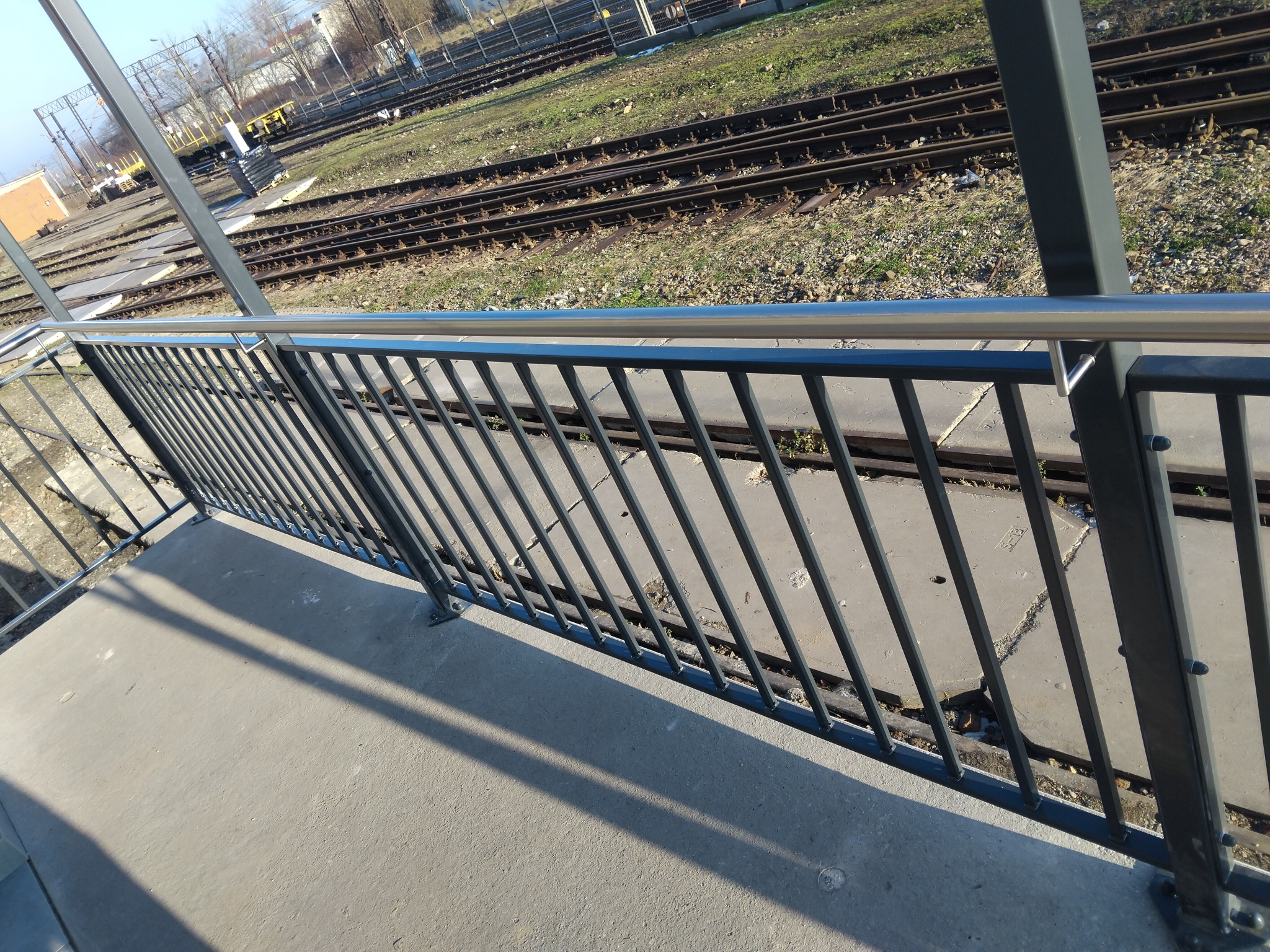 Metalowa balustrada z poziomą, nierdzewną poręczą na betonowym podeście, widok na tory kolejowe w tle.