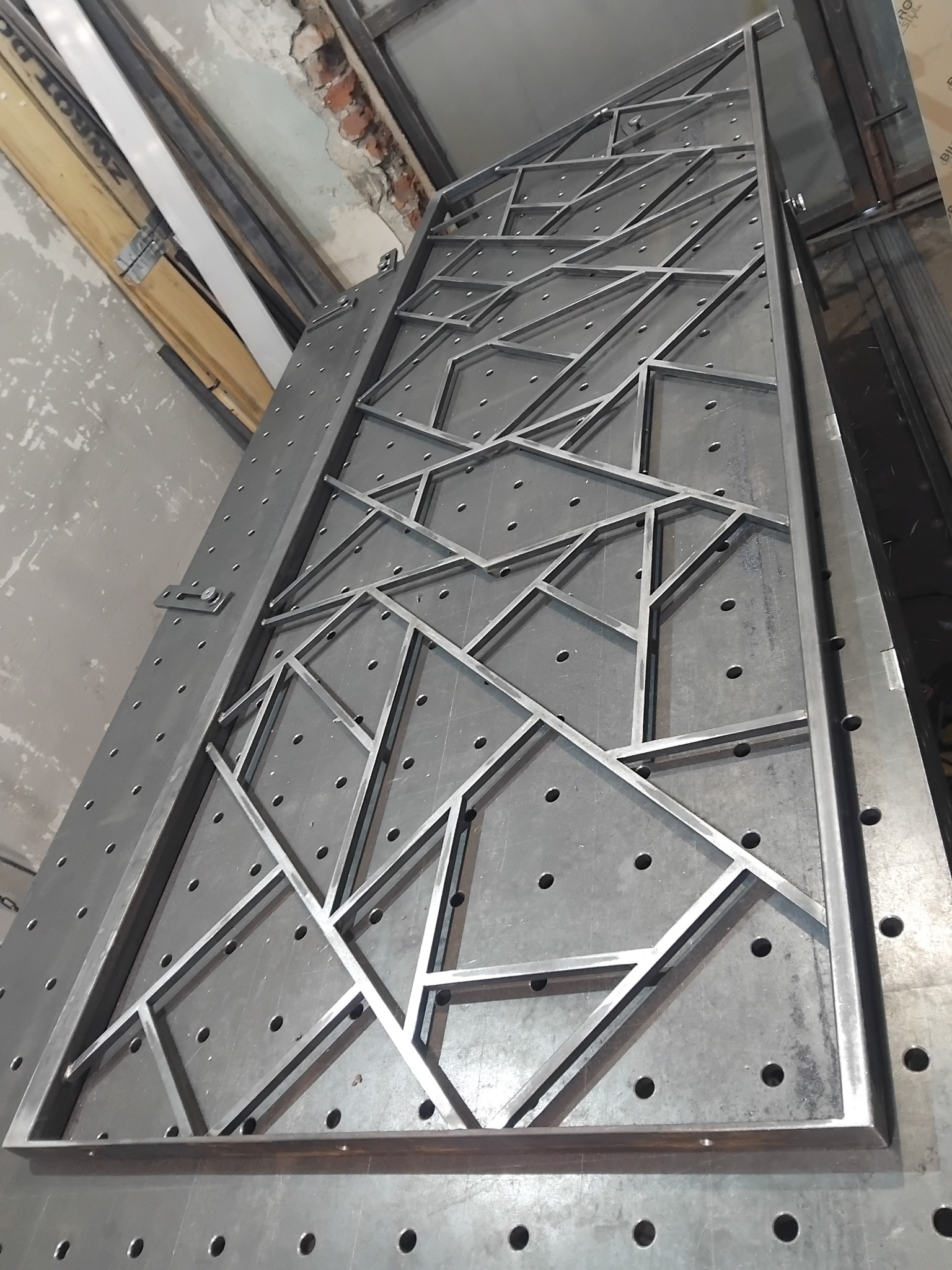 Metalowa konstrukcja z profili o nieregularnych kształtach geometrycznych, spawana na perforowanym stole spawalniczym, widoczne ślady obróbki i surowy stan wykończenia.