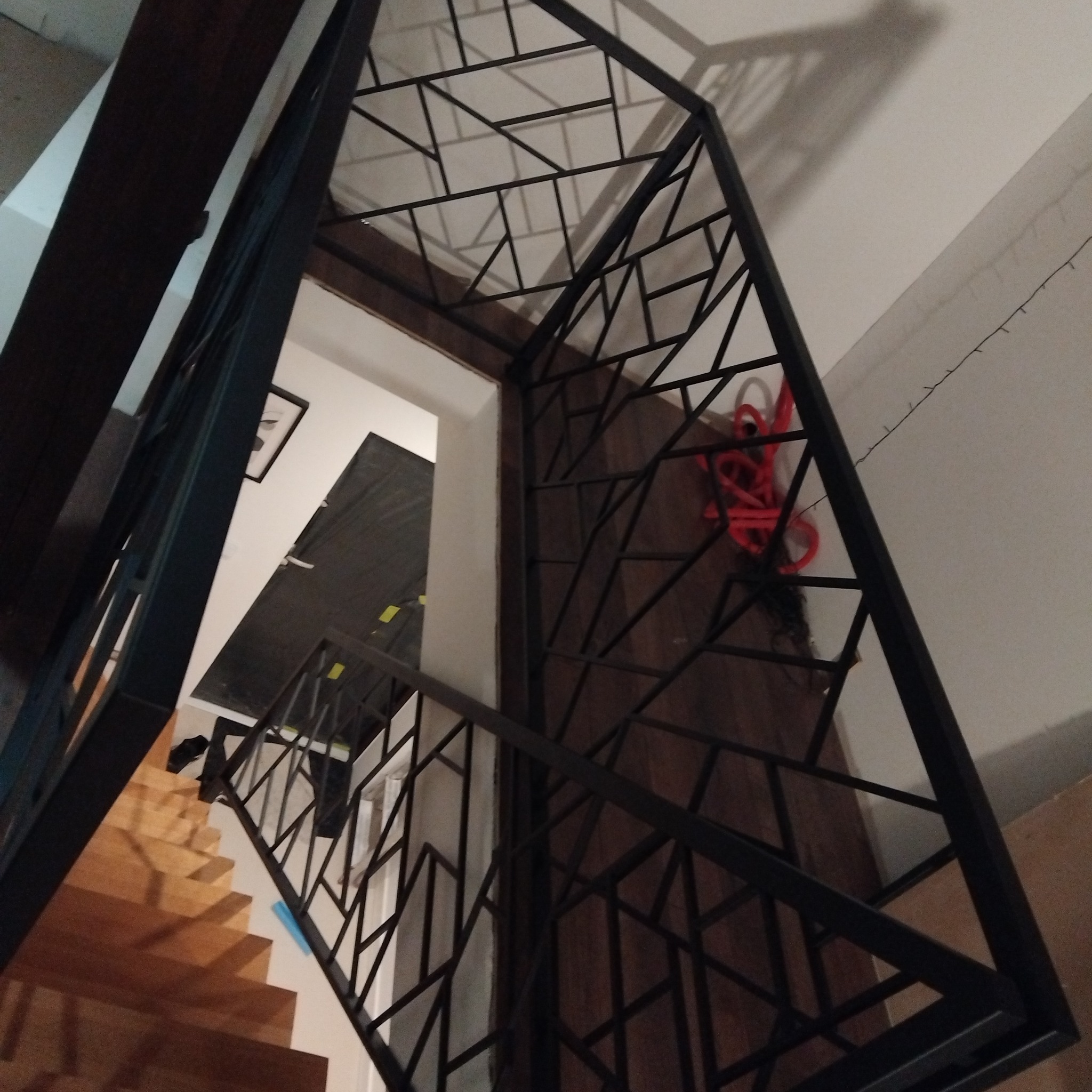 Metalowa konstrukcja schodów z geometrycznym wzorem, widok z dołu, schody drewniane w tle.