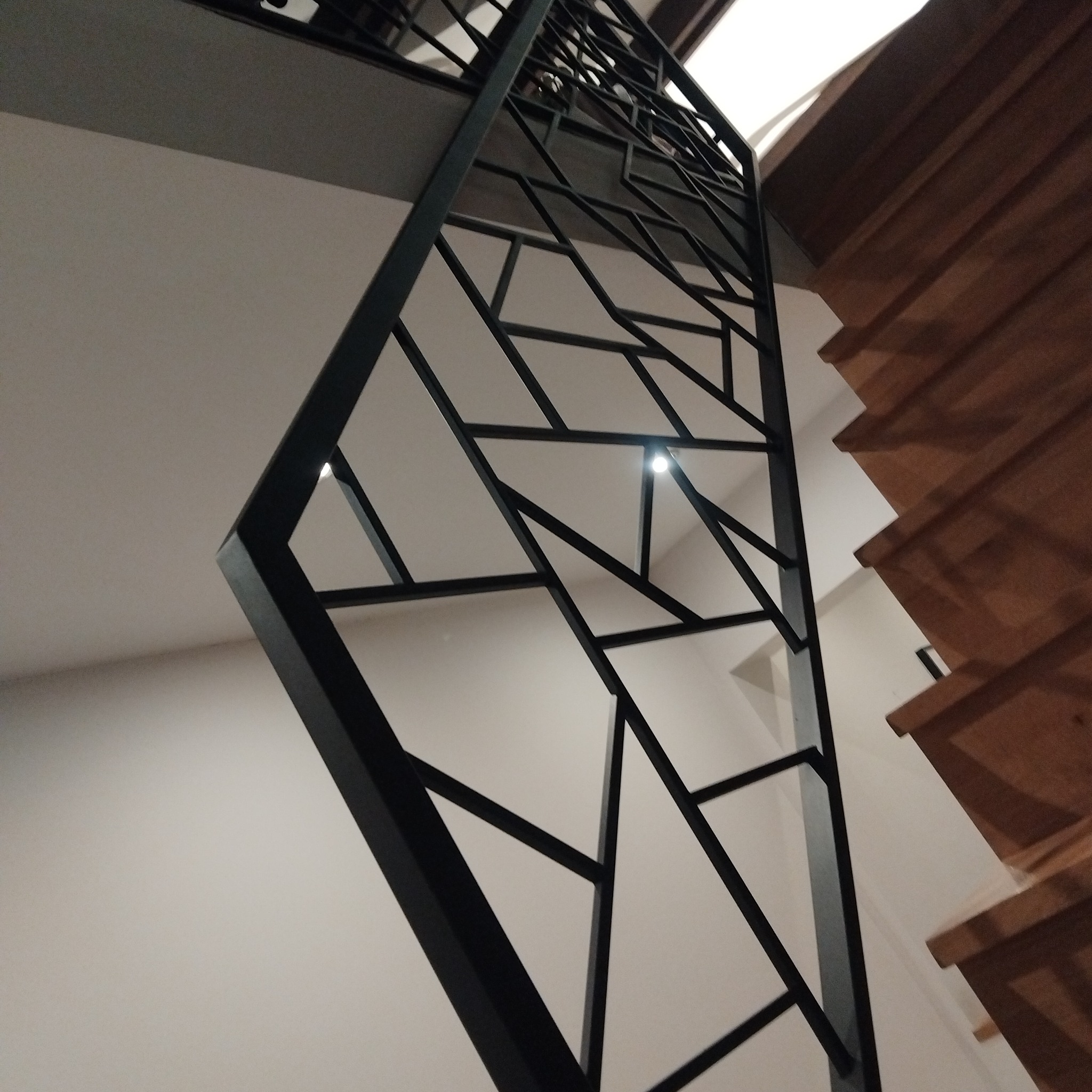 Nowoczesna, czarna, metalowa konstrukcja balustrady schodowej z geometrycznym wzorem, widziana z dołu, sąsiadująca z drewnianymi stopniami schodów.