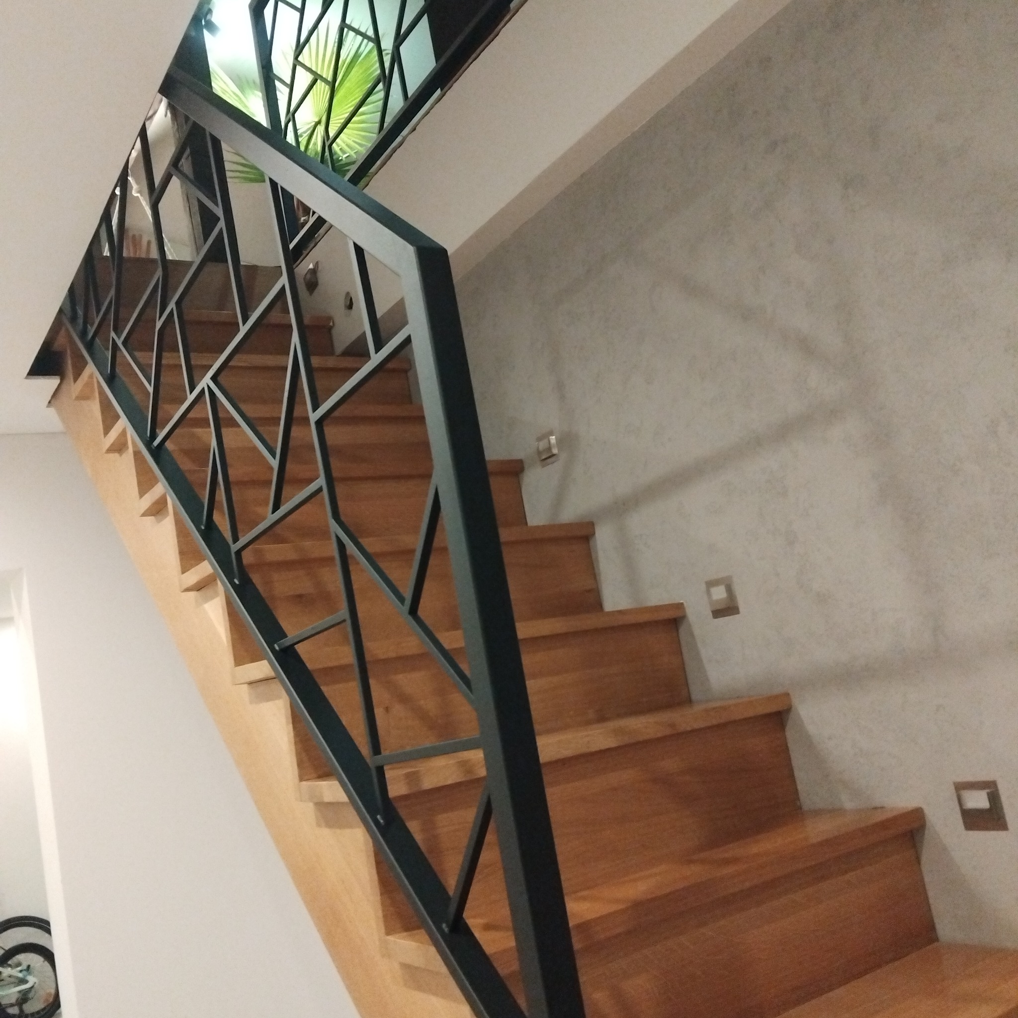 Drewniane schody z nowoczesną, geometryczną balustradą w kolorze grafitowym, widok z dołu, w tle ściana z fakturą i roślina doniczkowa.
