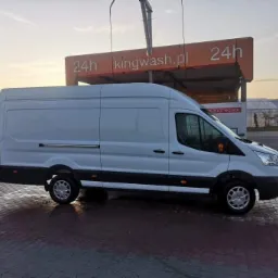 Biały bus dostawczy marki Ford zaparkowany na mokrym brukowanym placu przed myjnią samochodową Kingwash.pl o zmierzchu.