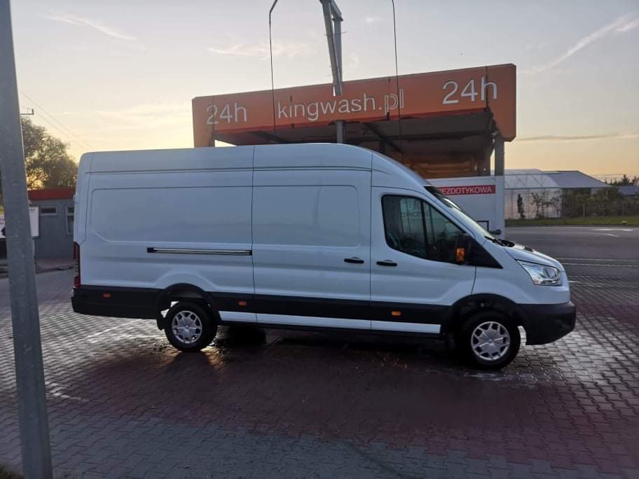 Biały bus dostawczy marki Ford zaparkowany na mokrym brukowanym placu przed myjnią samochodową Kingwash.pl o zmierzchu.