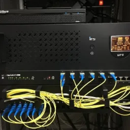 Czarna szafa rack z zamontowanym UPS z wyświetlaczem i panelem krosowniczym z podłączonymi kablami światłowodowymi oraz kablem ethernet. Widoczne diody sygnalizacyjne na panelu krosowniczym.