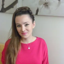 Portret kobiety z Lęborka, uśmiechniętej, z upiętymi włosami z warkoczem i wyrazistym makijażem oczu, w różowej bluzce i srebrnym naszyjniku z sercem.