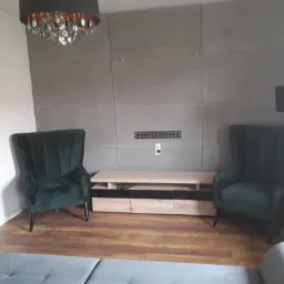 Pokój dzienny z szarą ścianą z paneli, dwoma zielonymi fotelami typu uszak, lampą podłogową oraz szafką RTV z jasnego drewna.