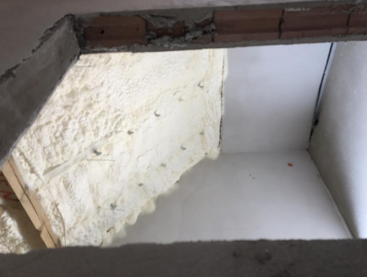 Wnętrze poddasza z nałożoną warstwą izolacji natryskowej, widoczne metalowe kotwy mocujące, surowy mur w górnej części kadru.