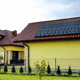 Żółty dom jednorodzinny z brązowym dachem i panelami fotowoltaicznymi na części dachu, na trawniku stoi biały samochód dostawczy z logo firmy instalującej fotowoltaikę.