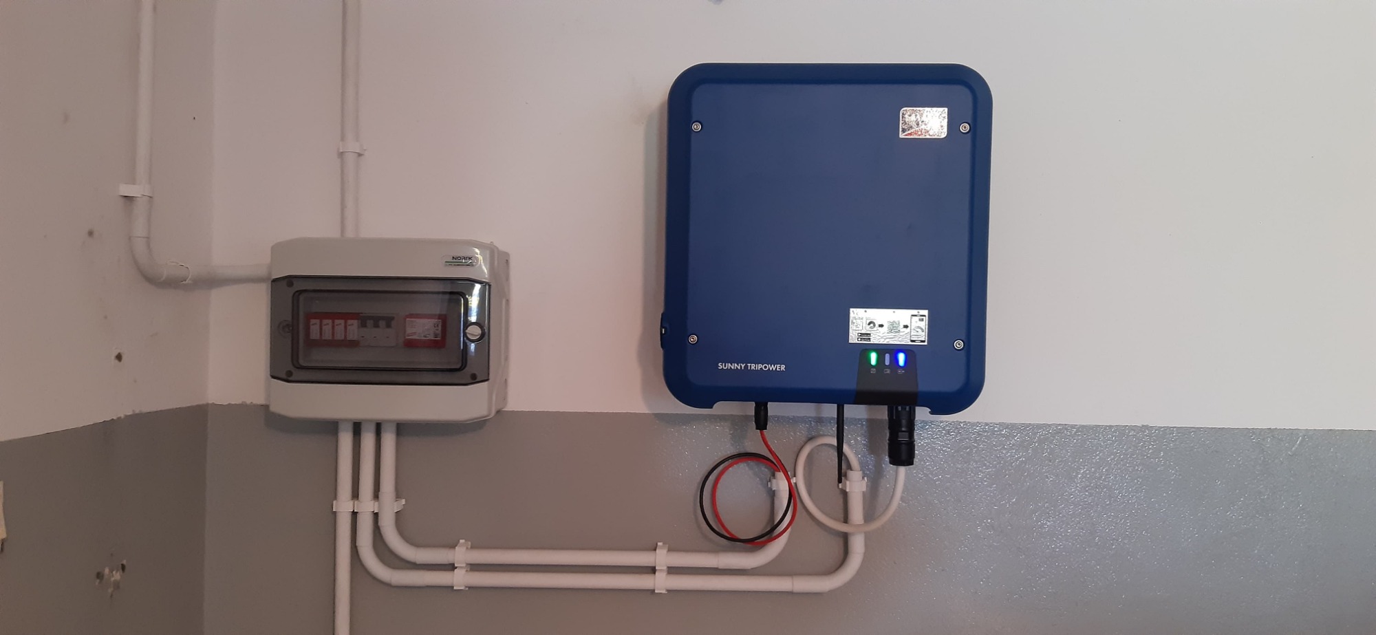 Niebieski inwerter Sunny Tripower zamontowany na ścianie obok skrzynki z bezpiecznikami, widoczne przewody i rury instalacyjne.