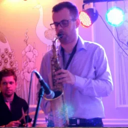 Saksofonista w okularach gra na saksofonie podczas występu na tle ściany z motywem ptaków i kwiatów, oświetlony kolorowymi reflektorami.