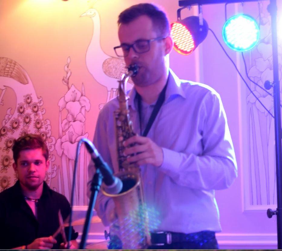 Saksofonista w okularach gra na saksofonie podczas występu na tle ściany z motywem ptaków i kwiatów, oświetlony kolorowymi reflektorami.