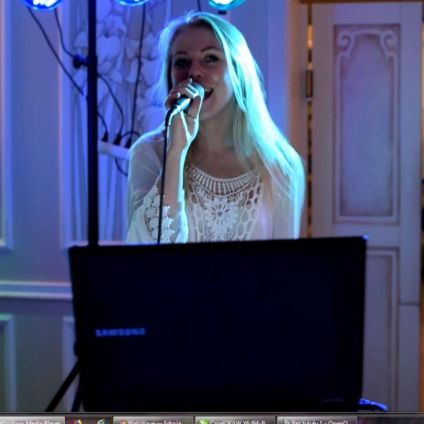 Blondynka śpiewa do mikrofonu przed laptopem Samsung na statywie, oświetlona niebieskim światłem, w tle dekoracyjne drzwi.