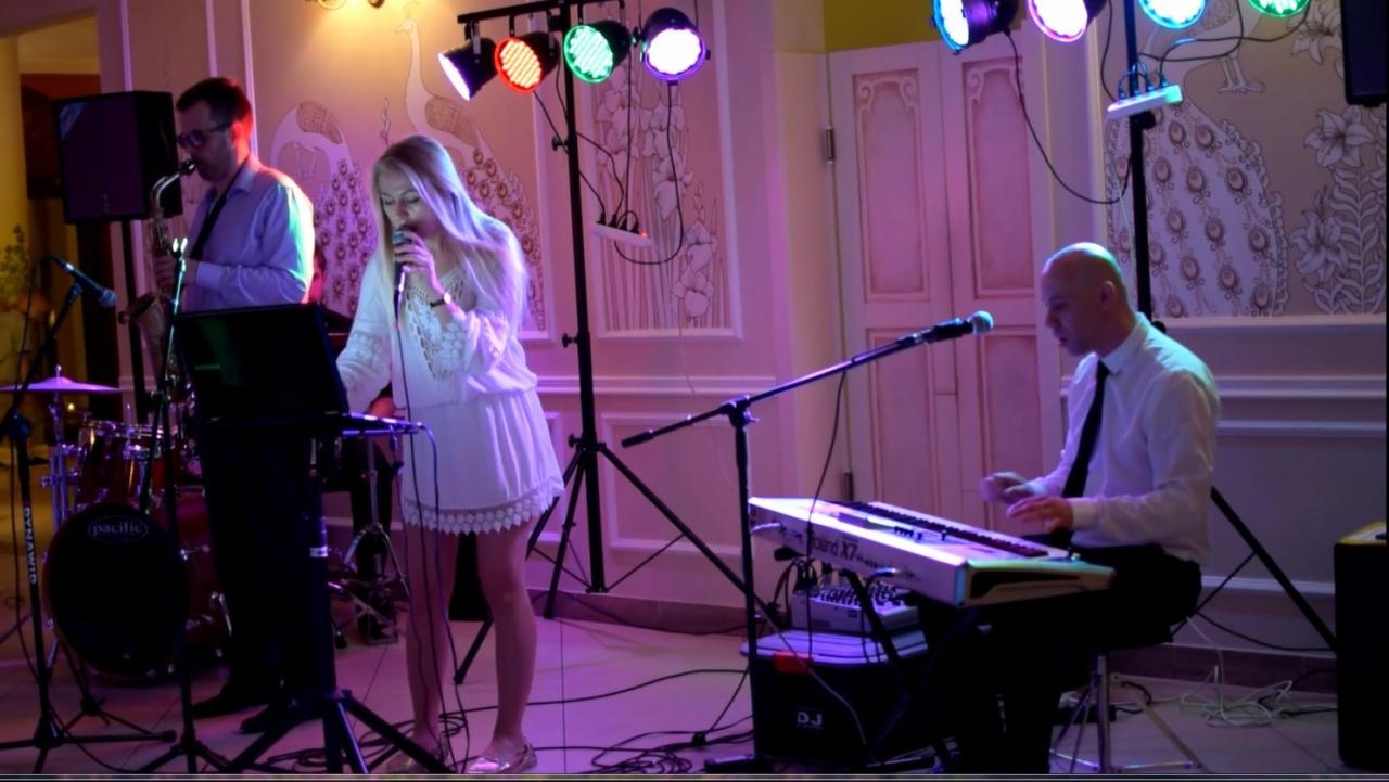 Zespół muzyczny w trakcie występu: wokalistka w białej sukience śpiewa do mikrofonu, saksofonista gra na saksofonie, klawiszowiec gra na keyboardzie Roland X7, oświetlenie sceniczne z kolorowymi...