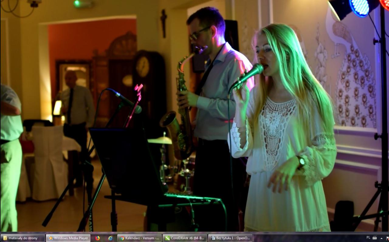 Wokalistka w białej sukience śpiewa do mikrofonu, obok saksofonista w okularach i koszuli, w tle dekoracja z motywem pawia i oświetlenie sceniczne.